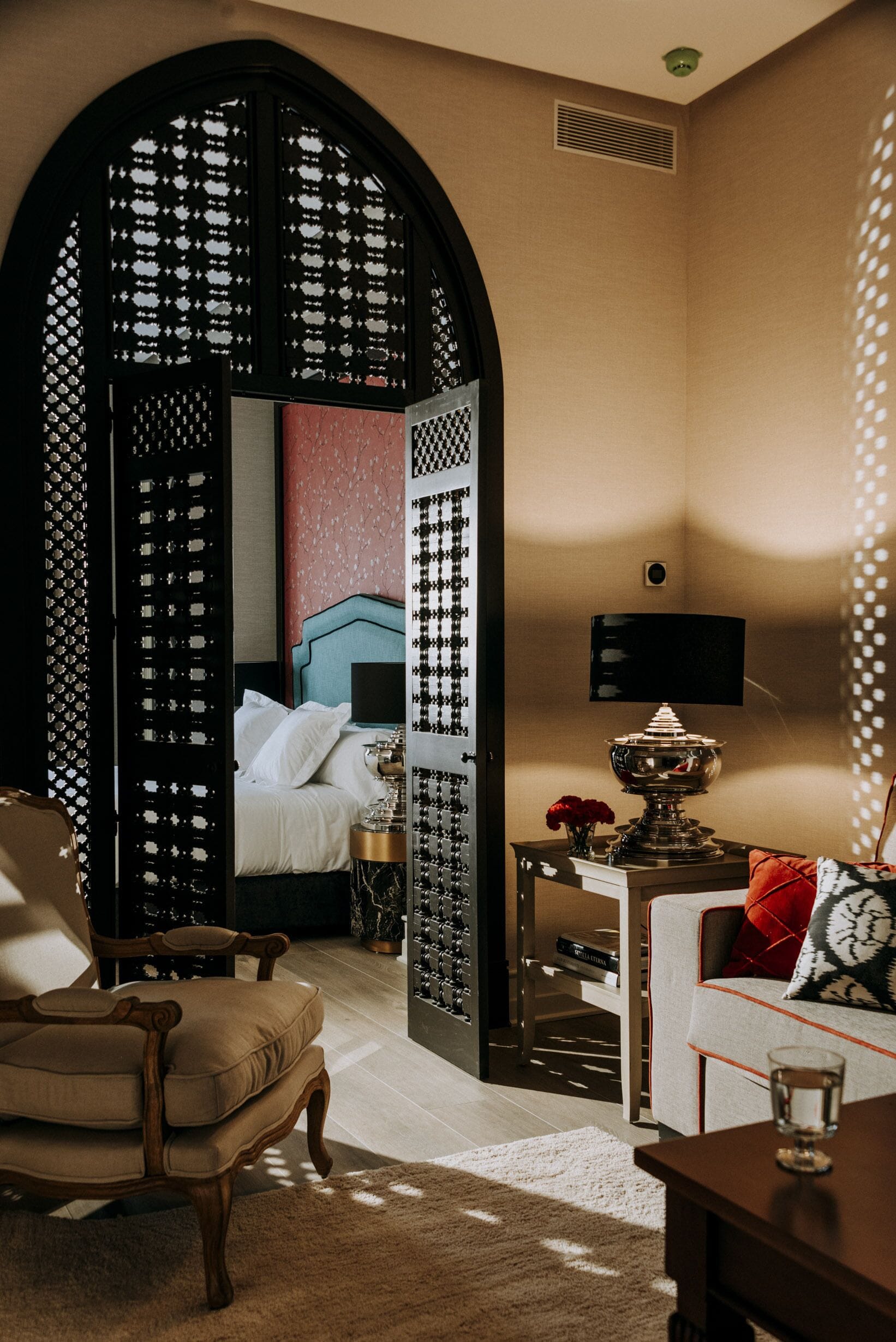 A hotel room with a large arched doorway leading to a bedroom, a sitting area with a sofa and armchair, a bedside table with a lamp, and a small table with a vase of flowers.