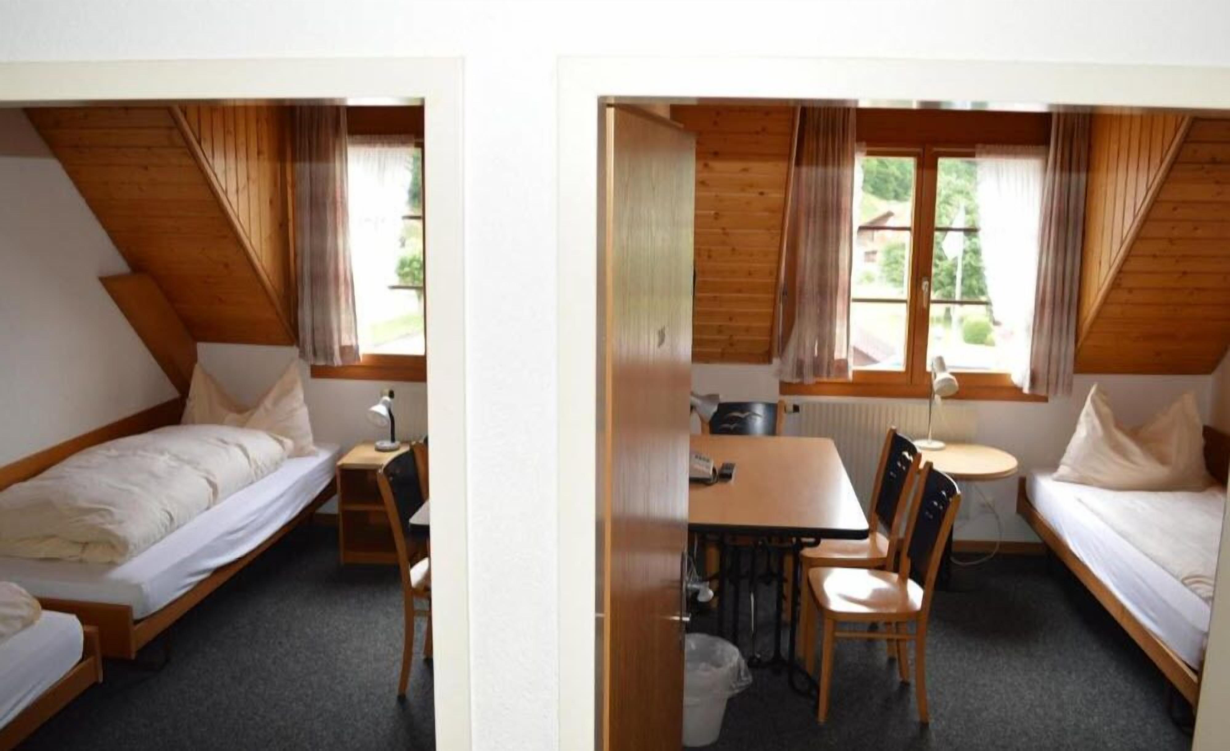 A small, cozy room with a bed, a desk, and a chair. There is a window with curtains, and the room has a sloped ceiling.
