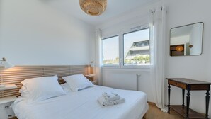 A neatly made bed with white linens and towels, a bedside table with a lamp, a wooden nightstand, a window with white curtains, and a mirror on the wall. - L'Estrade 4* - Appartement 2 bedrooms with the view lake & garage (Annecy)