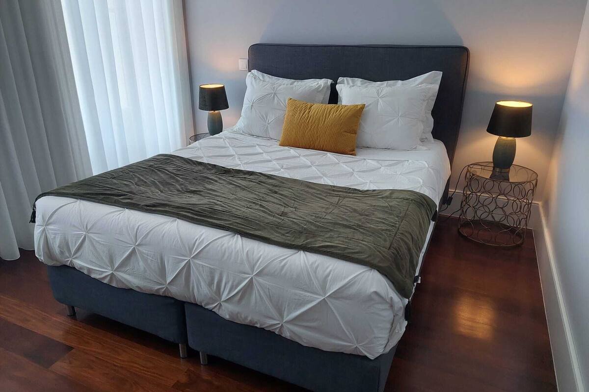 A neatly made bed with a dark headboard, white bedding, and a mustard-colored pillow. There are two bedside lamps on metal stands.