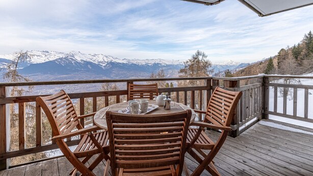 Une terrasse en bois avec une table ronde et des chaises, offrant une vue sur une chaîne de montagnes enneigées.