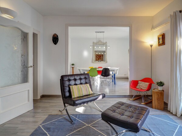 Una sala moderna con un sofá de cuero, un reposapiés a juego, un comedor con una mesa blanca y sillas coloridas, y una silla roja con una planta.