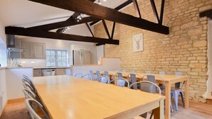 A spacious dining area with a long wooden table and metal chairs, a kitchen in the background, and exposed wooden beams on the ceiling. - The Cotswold Manor Wychwood, with Hot-Tub, Games Barn, 3 acres of Parkland (Witney)