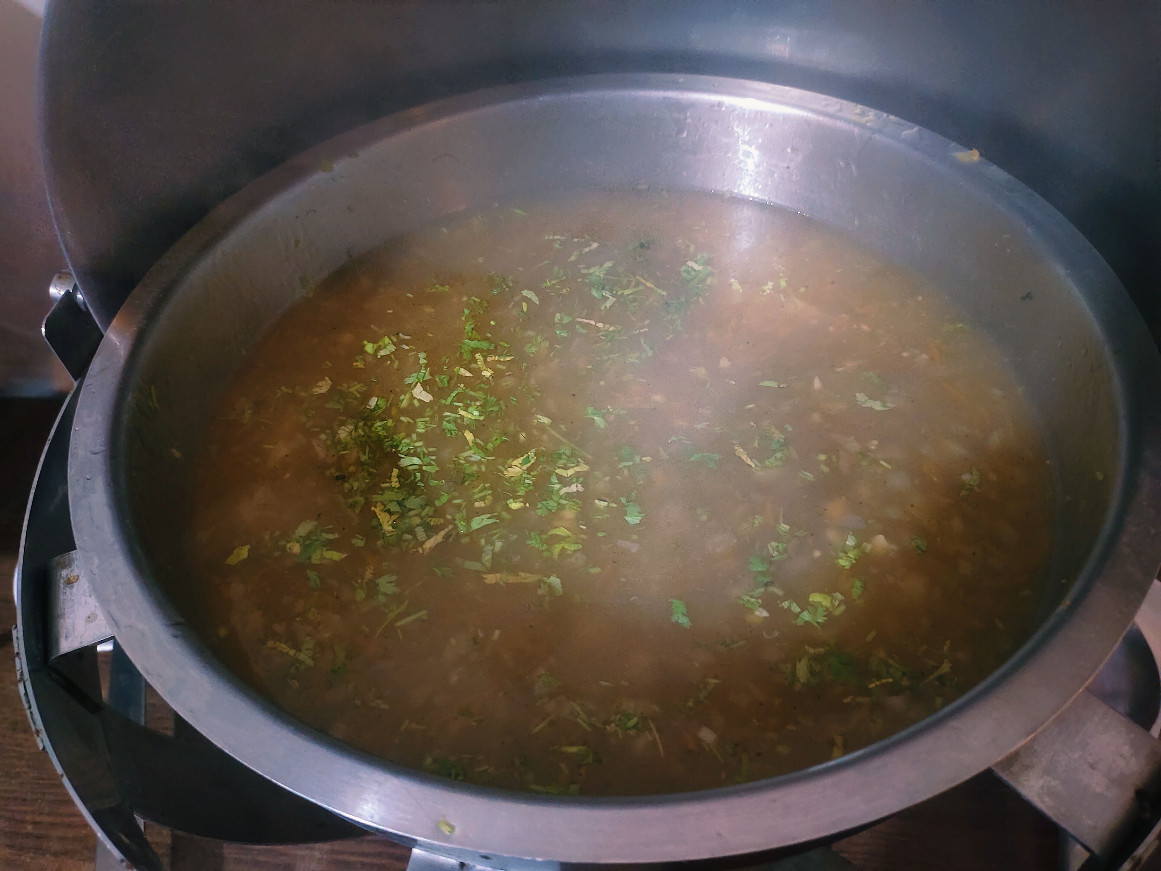 Uma panela de aço inoxidável com tampa, contendo um líquido transparente com ervas verdes, provavelmente uma sopa ou caldo, sobre um fogão.