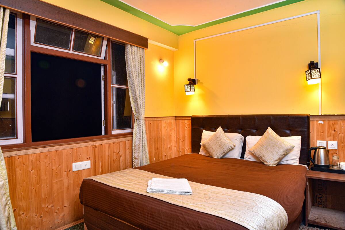 Une chambre dotée d’un grand lit, de lambris en bois, de deux lampes fixées au mur, d’une fenêtre avec des rideaux et d’une table de chevet avec une bouilloire.