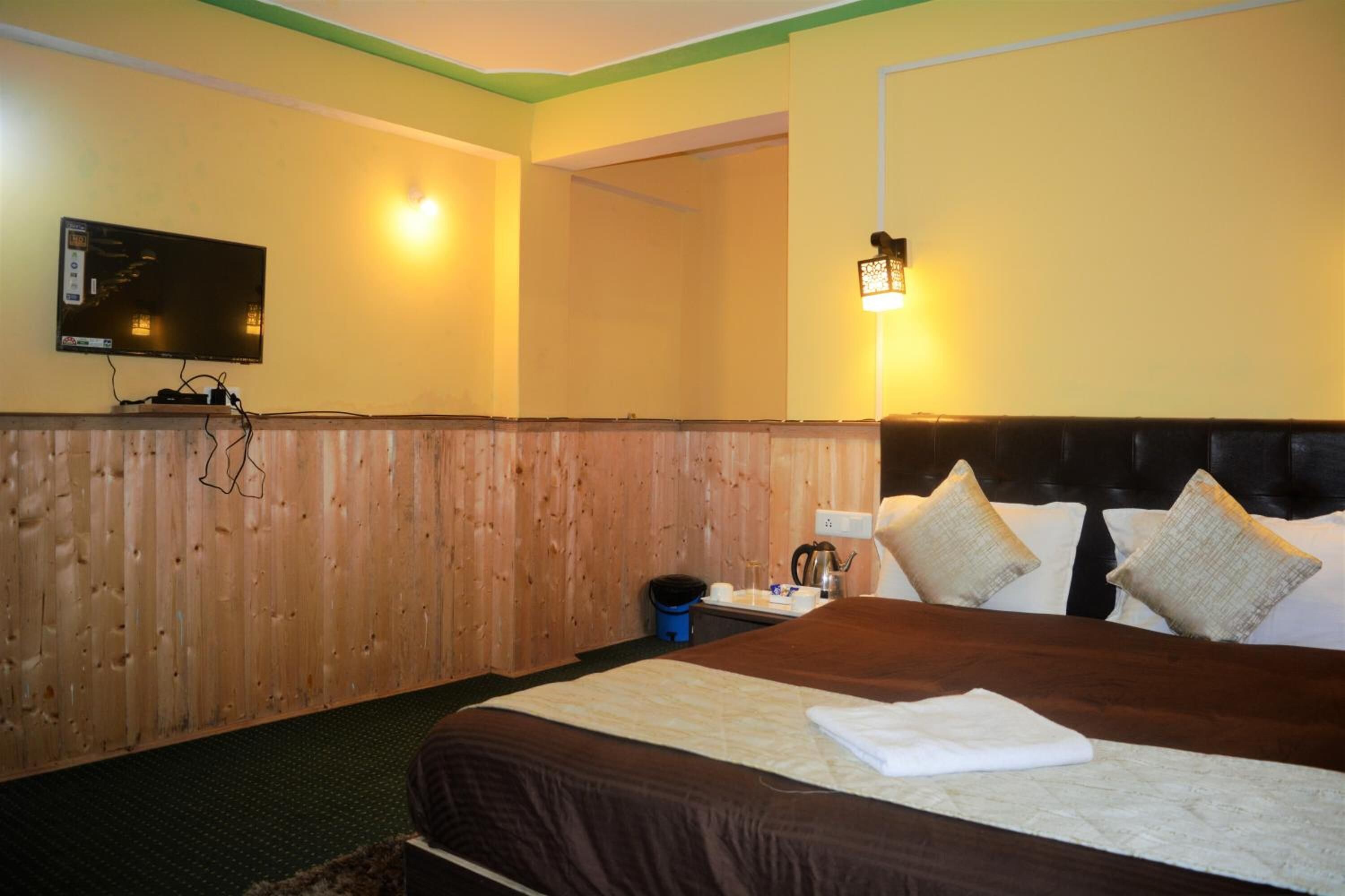 A hotel room with a wooden headboard, a flat-screen TV mounted on the wall, a bed with a brown bedspread and white pillows, and a small table with a kettle and cups.