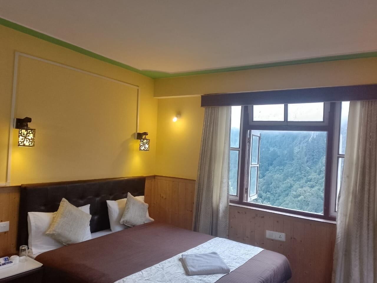 A hotel room with a large bed, two wall-mounted lamps, a window with a view of a forest, and a wooden headboard.