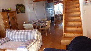 Een gezellige kamer met een houten trap, een eethoek met een tafel gedekt voor vier personen, en een bank met gestreepte kussens.