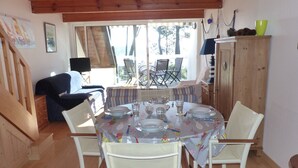 Een eetruimte met een ronde tafel voor vier personen, omringd door witte stoelen. Aan de rechterkant staat een houten kast en op de achtergrond een bank met een gestreept kussen.