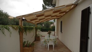 Una veranda riparata con tavolo e sedie, circondata da pareti bianche e travi di legno.