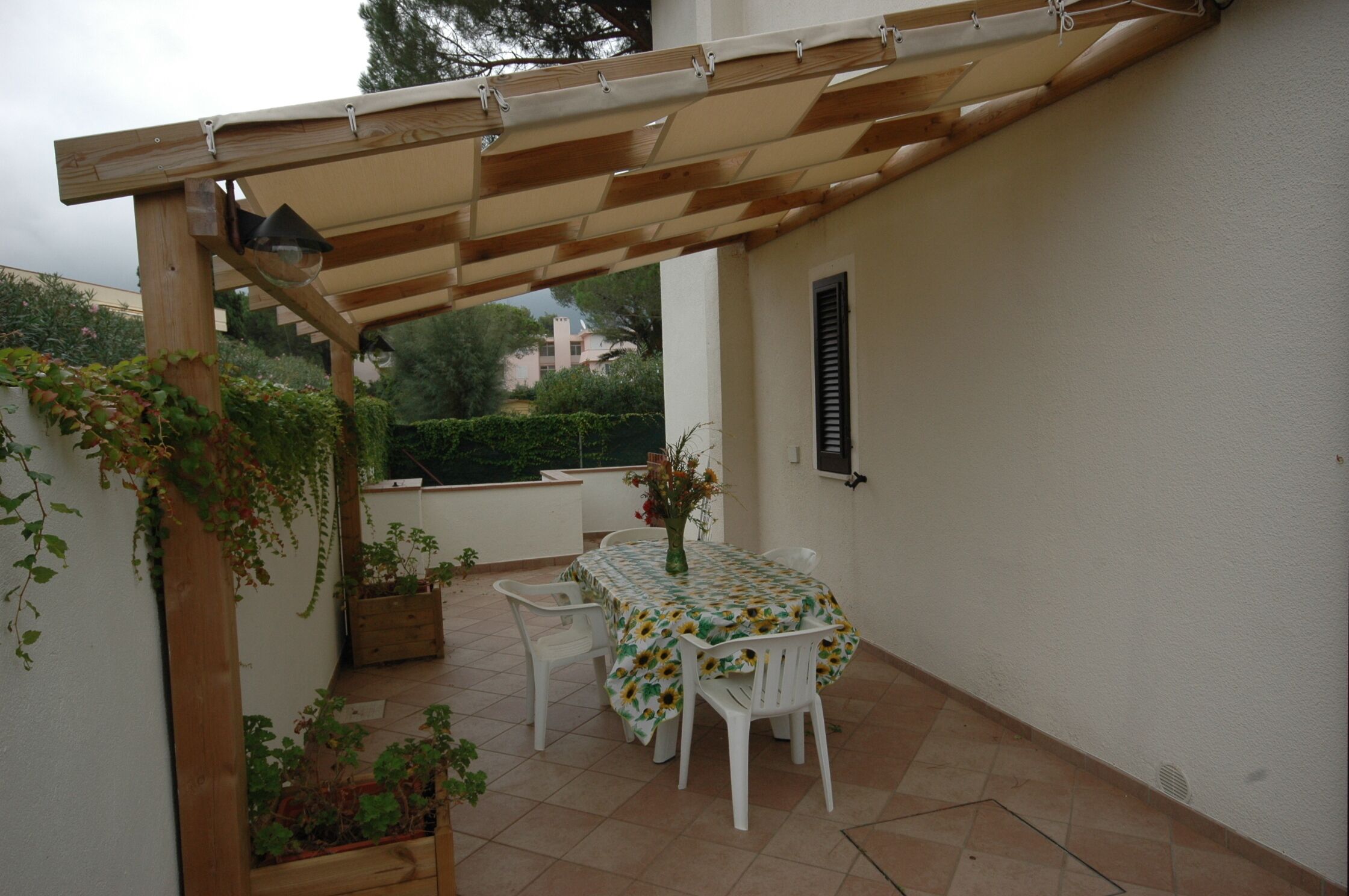Un espacio de patio cubierto con mesa y sillas, una pérgola de madera y plantas en macetas.