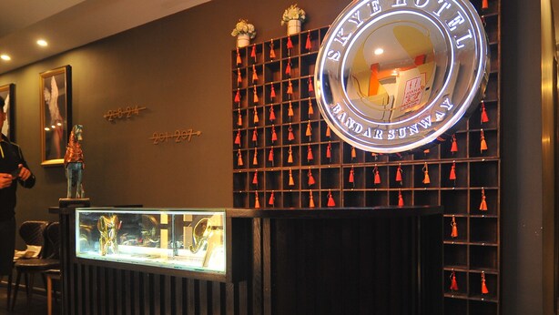 Een receptiebalie met een glazen vitrine, een neonreclame erboven en een muur met een raster van kleine rode decoraties.