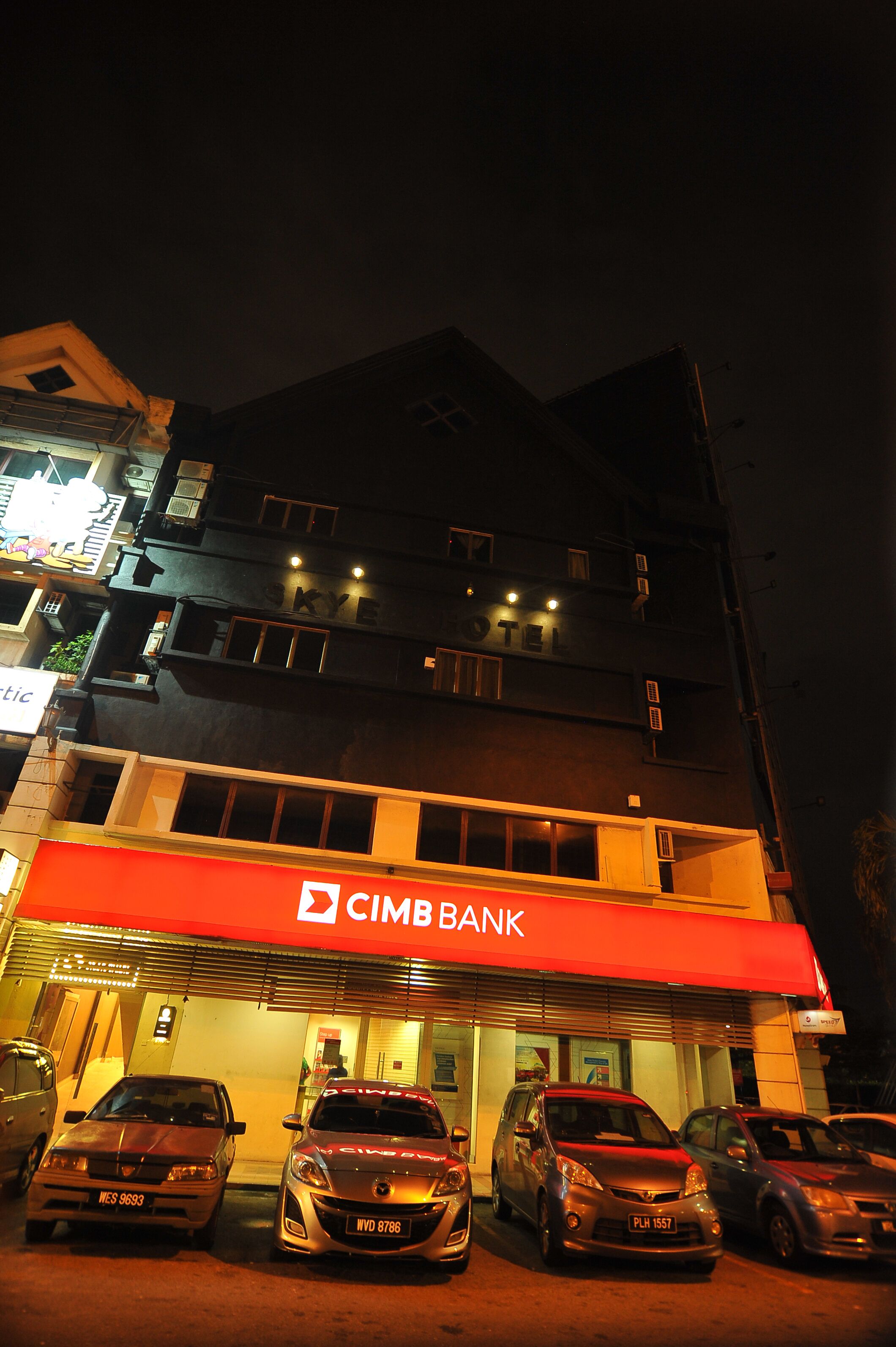 Un edificio de varios pisos con un toldo rojo que dice "CIMB BANK" y varios coches aparcados delante.