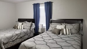 Habitación de hotel con dos camas individuales, una ventana con cortinas azules y una mesita de noche con lámpara.