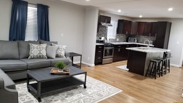 Una sala moderna con un sofá gris, una mesa de centro con una bandeja y una planta, y una zona de cocina con armarios oscuros, un microondas y una estufa.