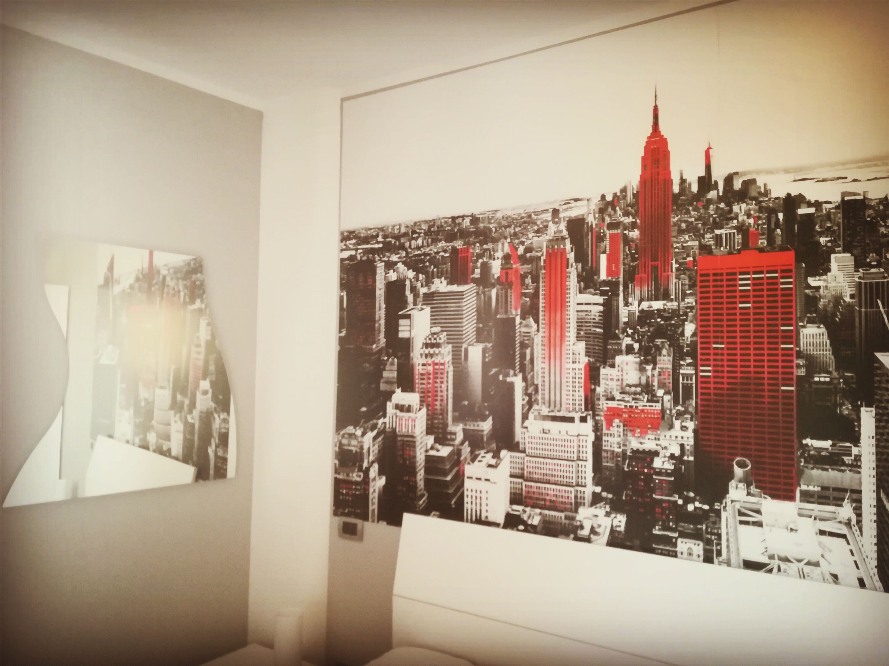 Une pièce avec une grande affiche représentant un paysage urbain accrochée au mur, un lit blanc et une vue partielle sur une fenêtre.