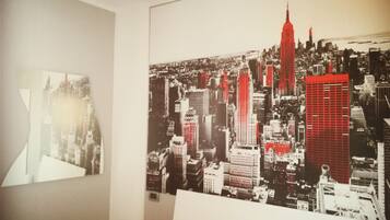 Une pièce avec une grande affiche représentant un paysage urbain accrochée au mur, un lit blanc et une vue partielle sur une fenêtre.