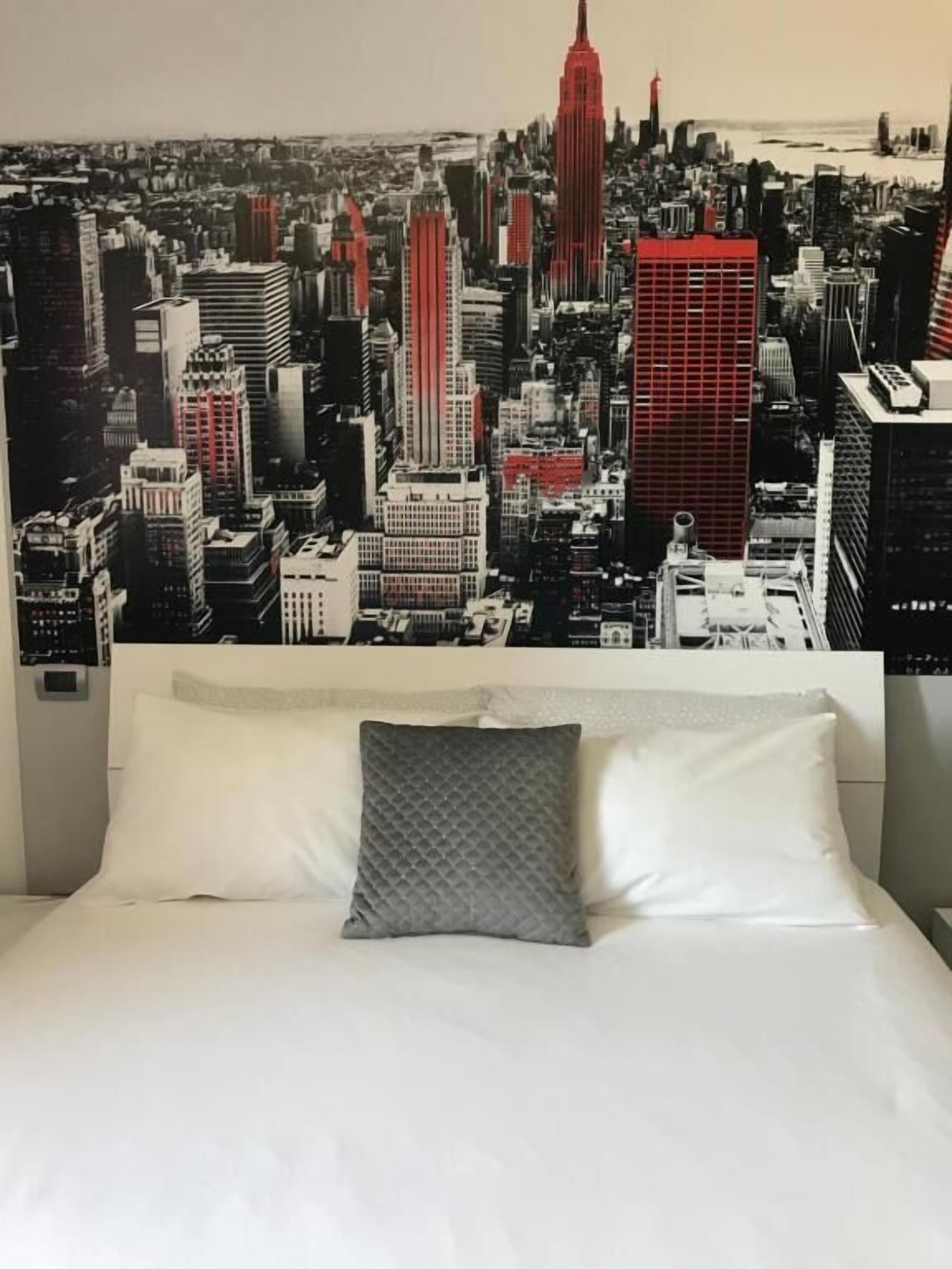 Een bed met wit beddengoed en een grijs kussen, voor een muur met een stadsgezichtsmural.