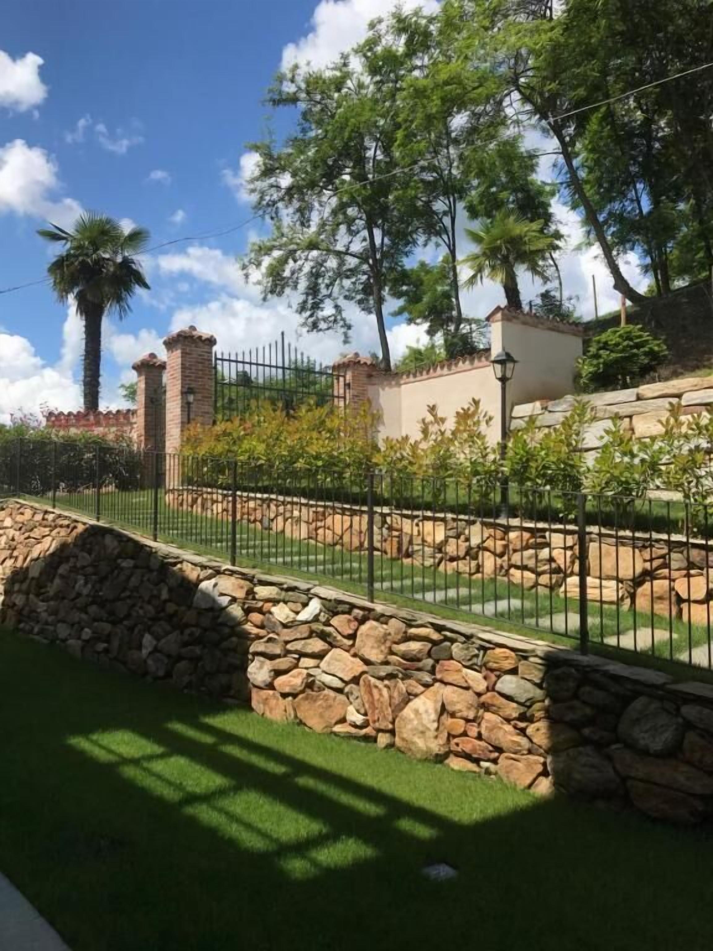Un'area giardino recintata con un muro in pietra, una palma e un edificio con tetto in tegole.