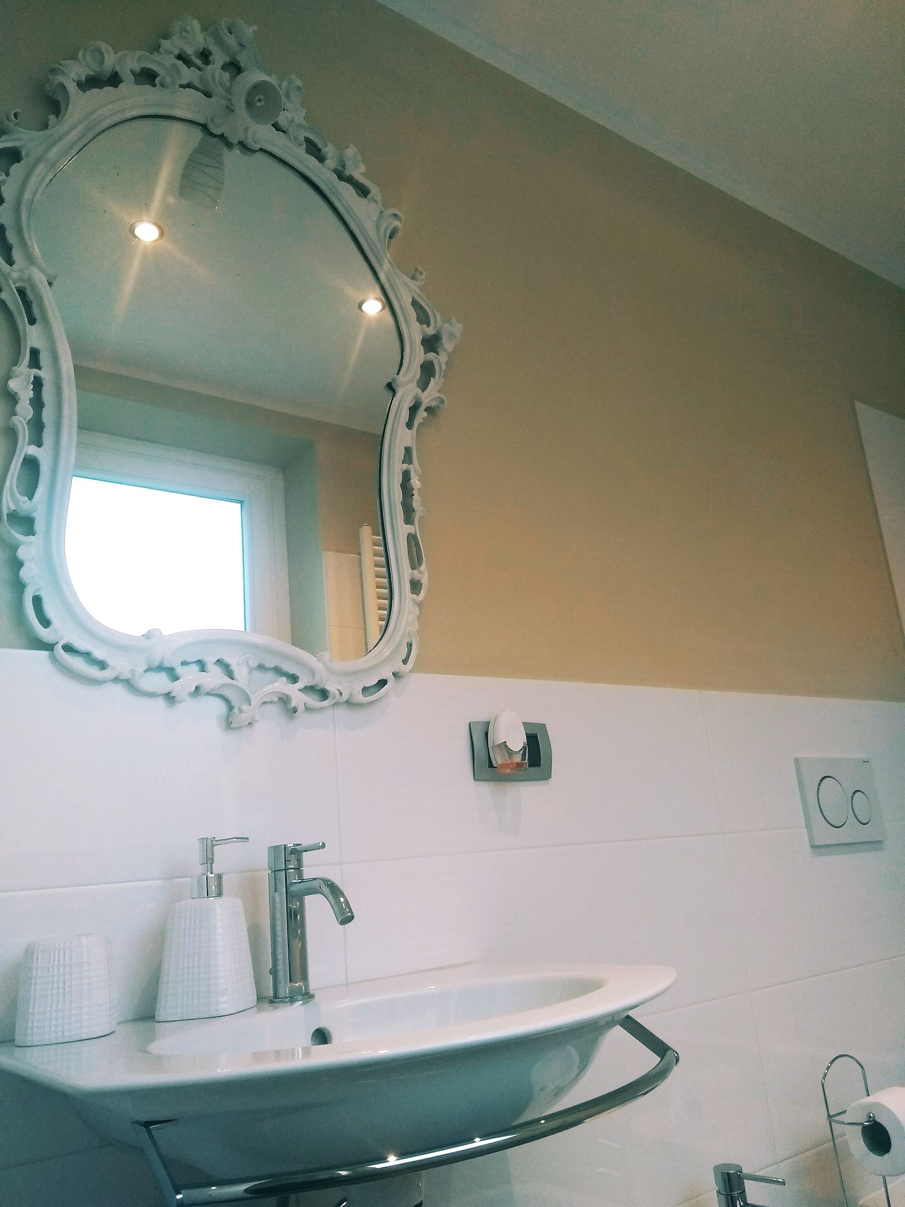 Un baño con un lavamanos blanco, un espejo con detalles ornamentales y un ventanal que proporciona luz natural.