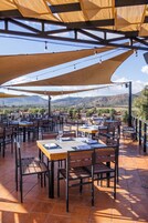 Un área de comedor al aire libre con varias mesas y sillas, protegida por grandes toldos, con vistas a un paisaje montañoso.