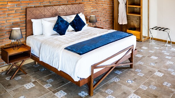Una cama bien hecha con estructura de madera, dos mesitas de noche con lámparas, una manta azul y un fondo de pared de ladrillo.