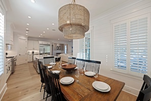 A dining area with a wooden table set for six, surrounded by black chairs, a woven chandelier, and white paneled walls.