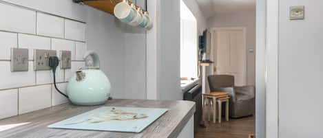 Una cocina con encimera de madera, tetera y cafetera. Hay armarios sobre la encimera y una silla al fondo.