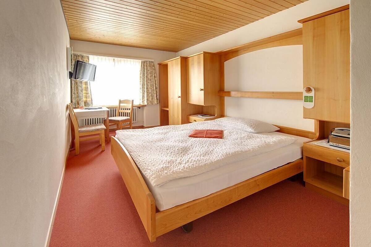 Quarto com cama de madeira, escrivaninha com cadeira, televisão e janela com cortinas.