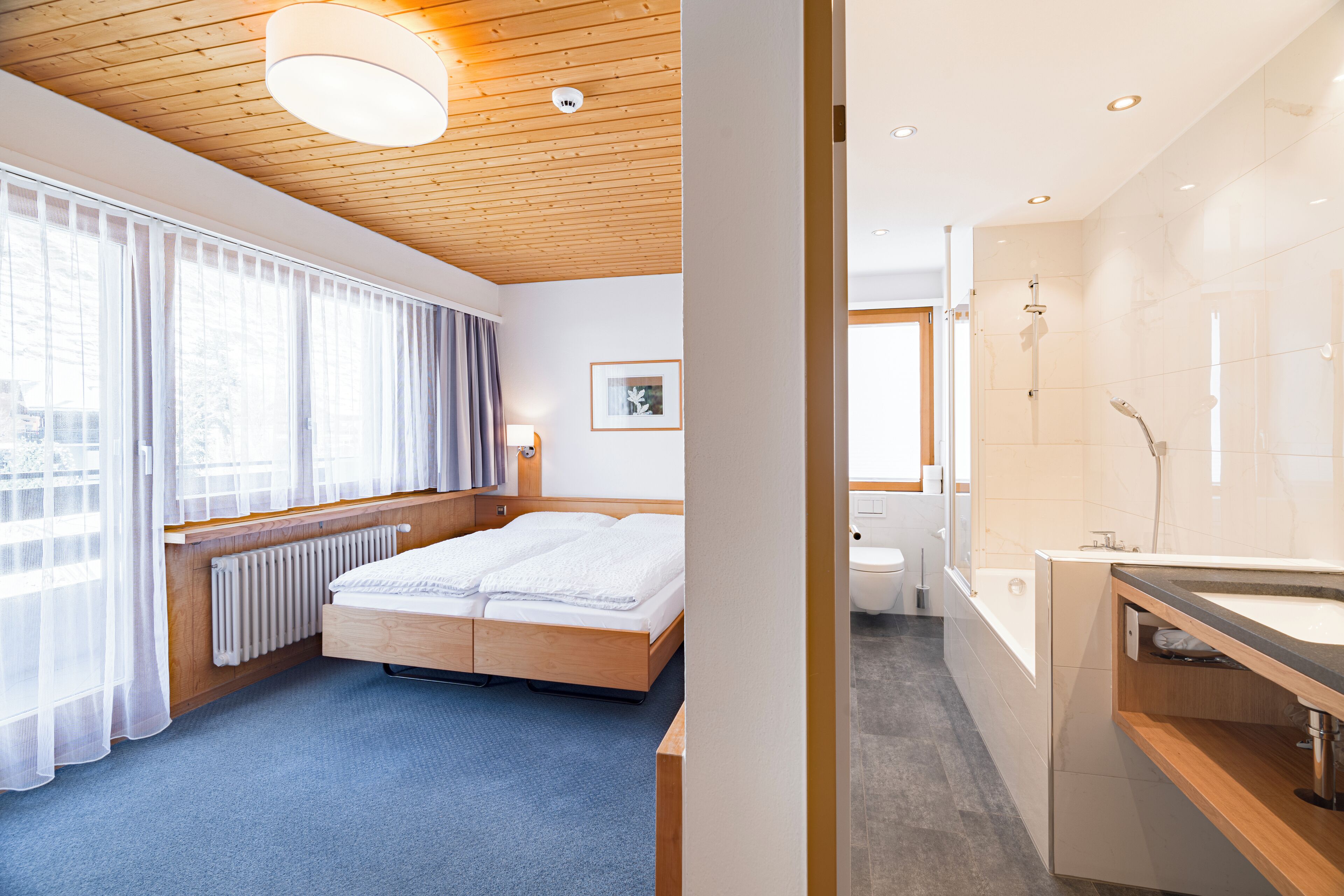 Une chambre d’hôtel avec un plafond en bois, une grande fenêtre avec des rideaux, un lit, un radiateur et une salle de bain équipée d’une baignoire et d’un lavabo.