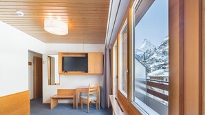 A hotel room with a wooden ceiling, a bed, a desk with a chair, a television, and a window with a view of snow-covered mountains. - Hotel Parnass (Zermatt)