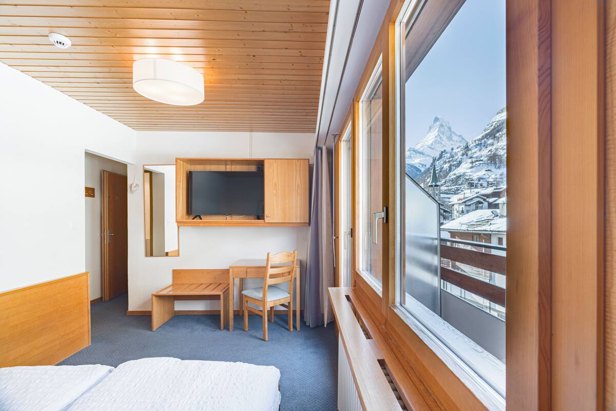 Quarto de hotel com teto de madeira, uma cama, escrivaninha com cadeira, televisão e janela com vista para montanhas cobertas de neve.
