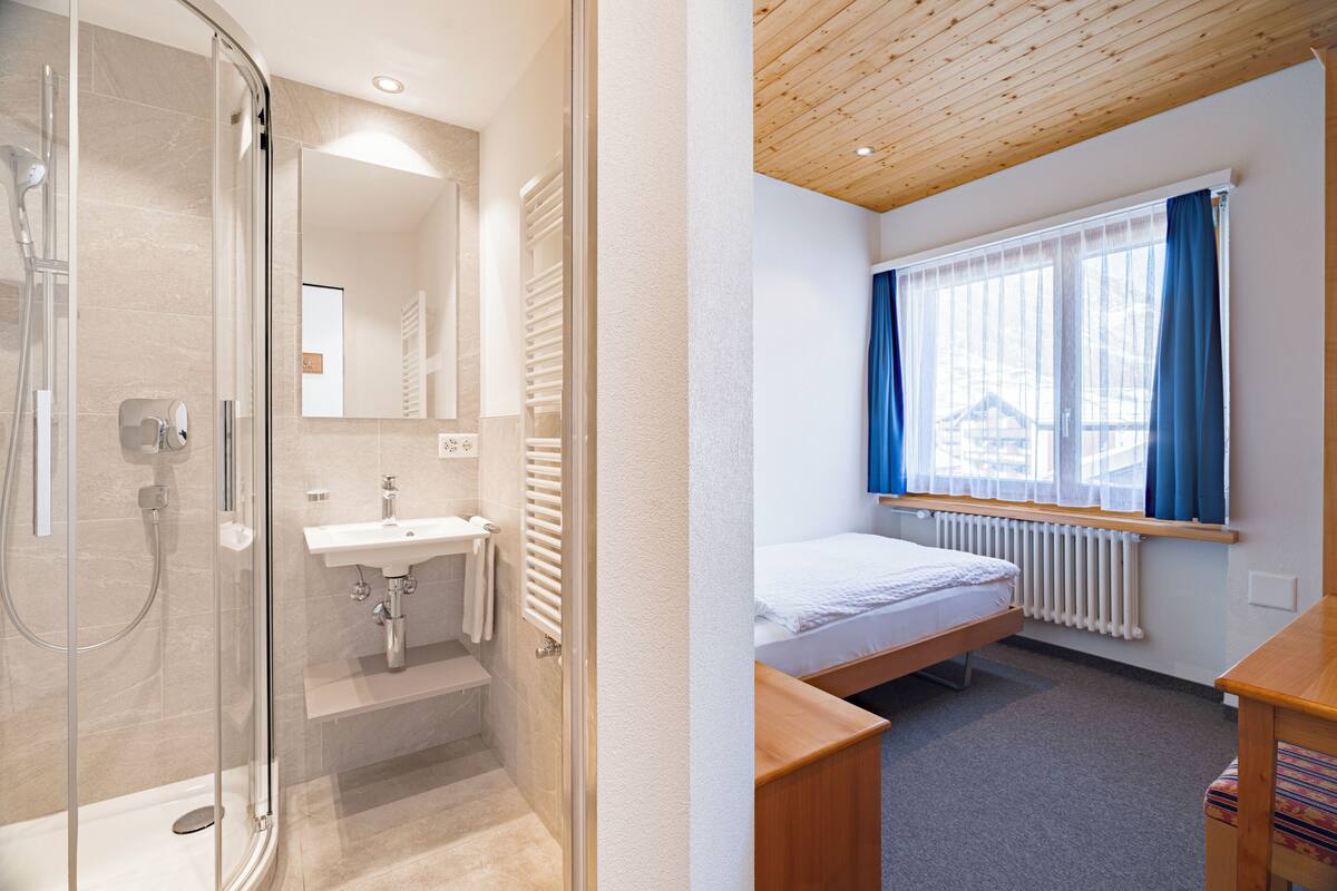 Um banheiro moderno com box de vidro, pia e vaso sanitário. Há uma cama com cabeceira de madeira e uma janela com cortinas azuis.