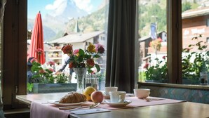 A table with a croissant, a glass of water, and a cup, set against a window with a view of mountains and a red umbrella outside. - Hotel Parnass (Zermatt)