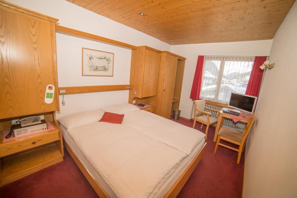 Une chambre à coucher comprenant un lit, une armoire en bois, un bureau, une chaise et une fenêtre avec des rideaux.