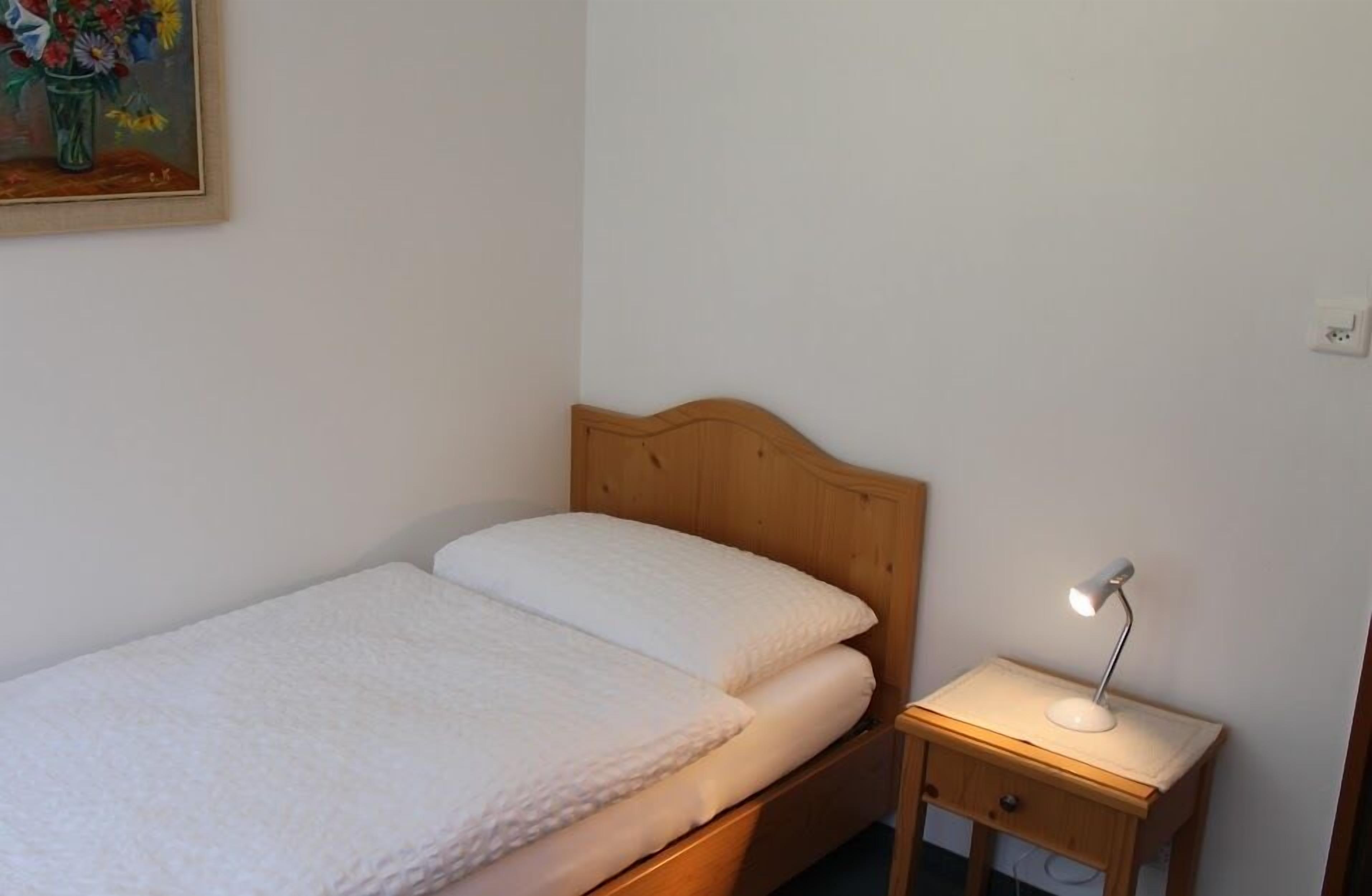 Una cama individual con cabecera de madera, una mesita de noche con lámpara y un cajón pequeño.