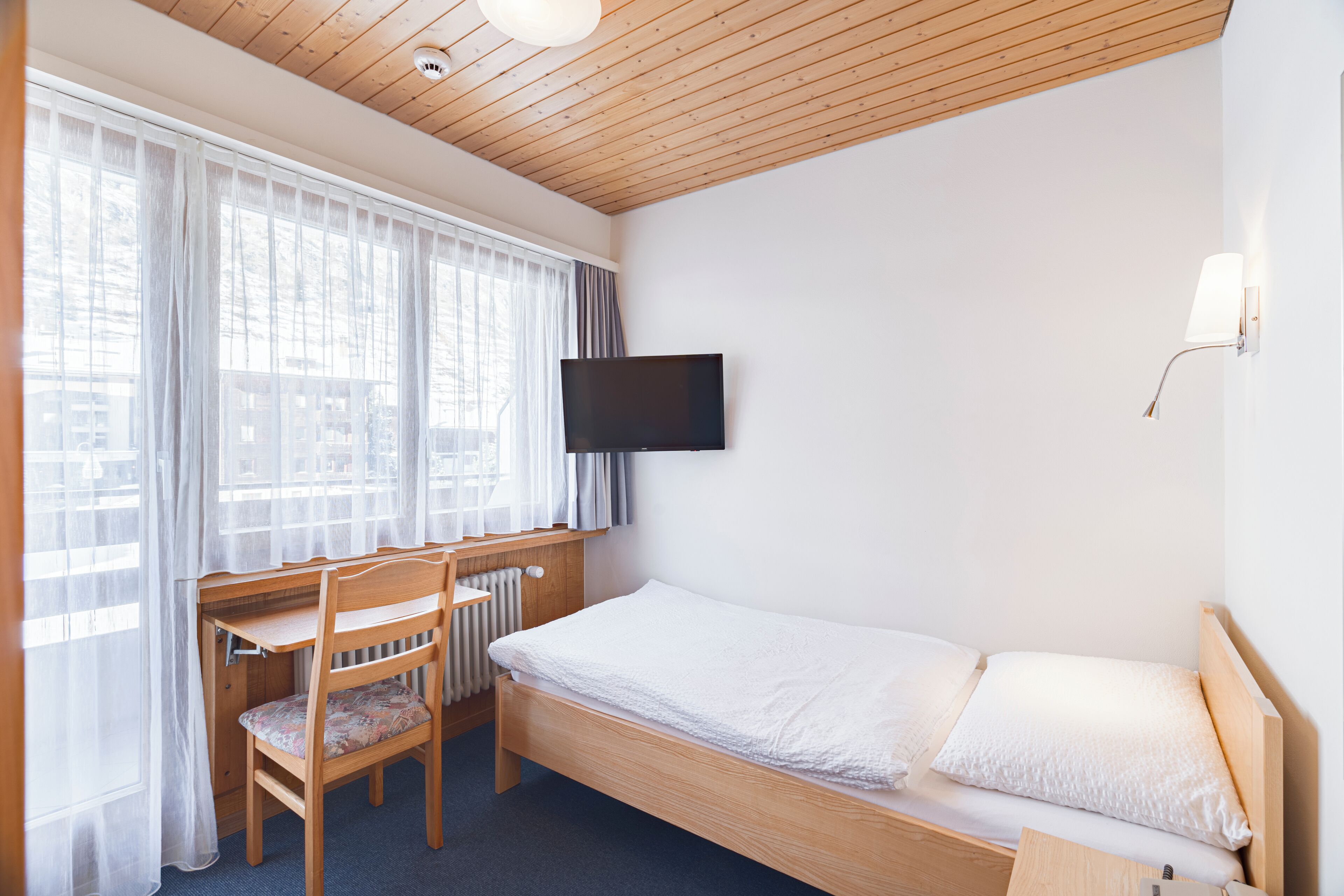 A bedroom with a wooden bunk bed, a desk with a chair, a television mounted on the wall, and a window with sheer curtains.