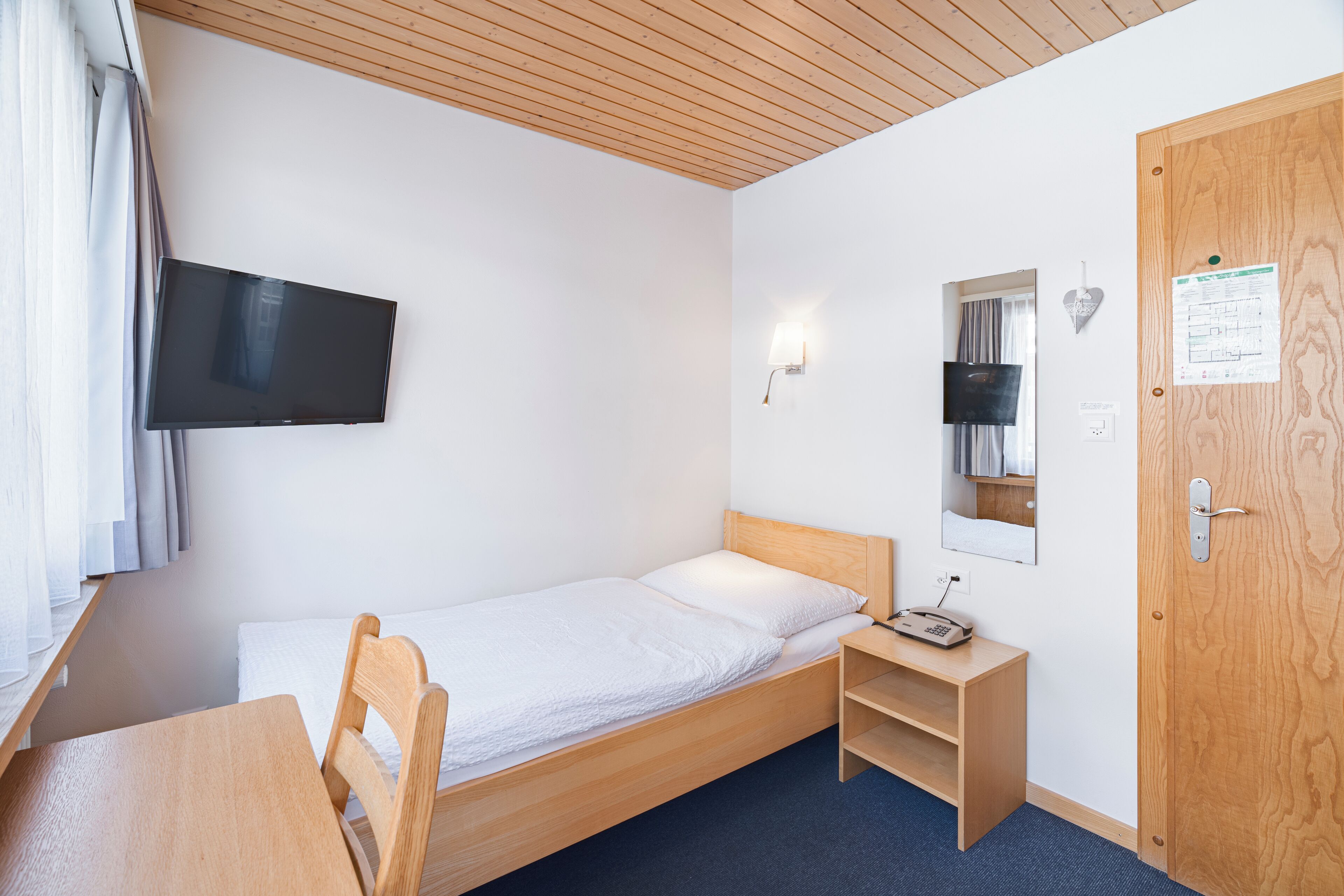 A single-bed room with a wooden headboard, a small bedside table with a lamp, a mounted TV, a mirror, and a door with a wooden panel.