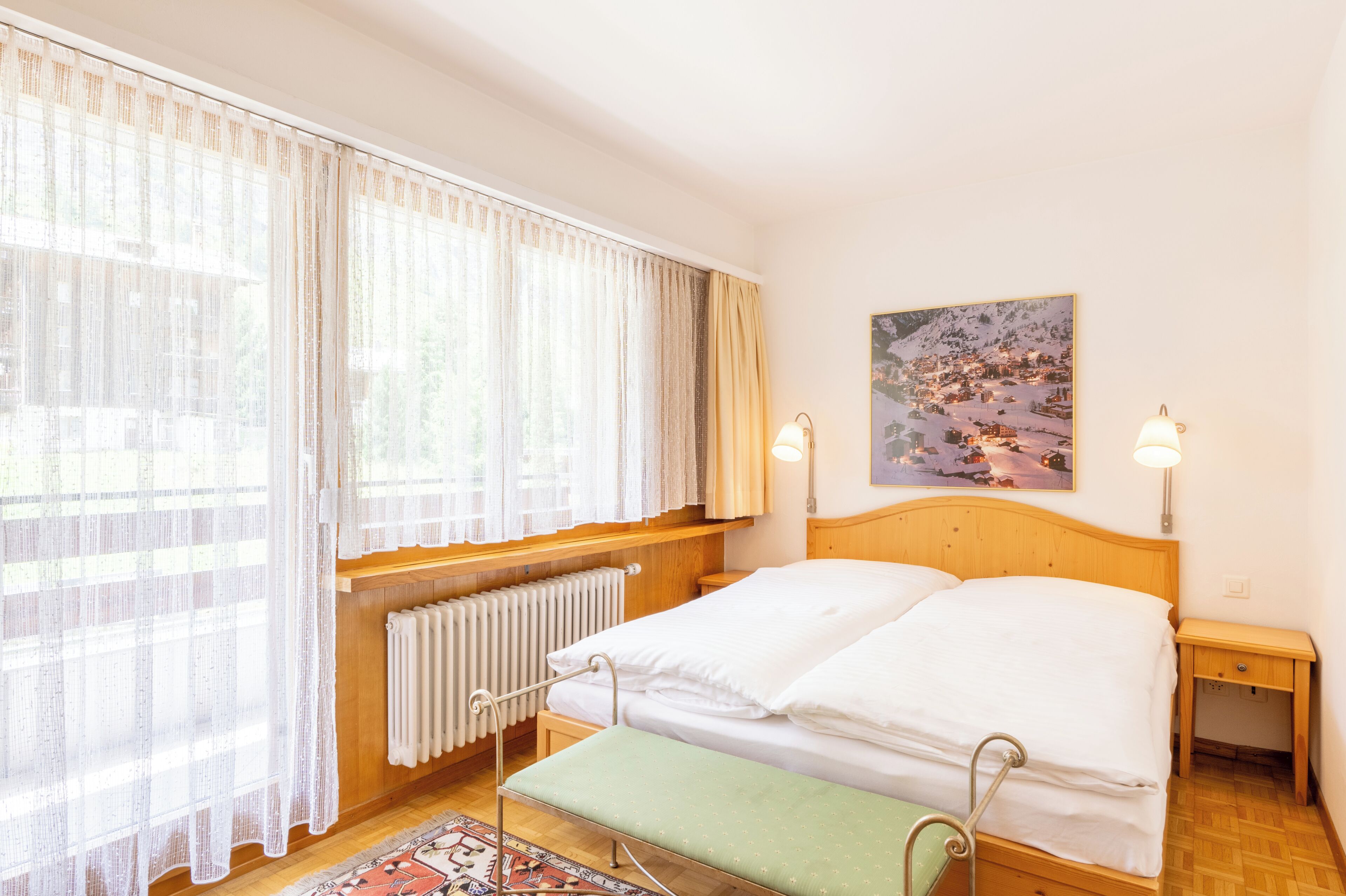 A bedroom with a bed, bedside tables, a radiator, a window with curtains, and a painting on the wall.