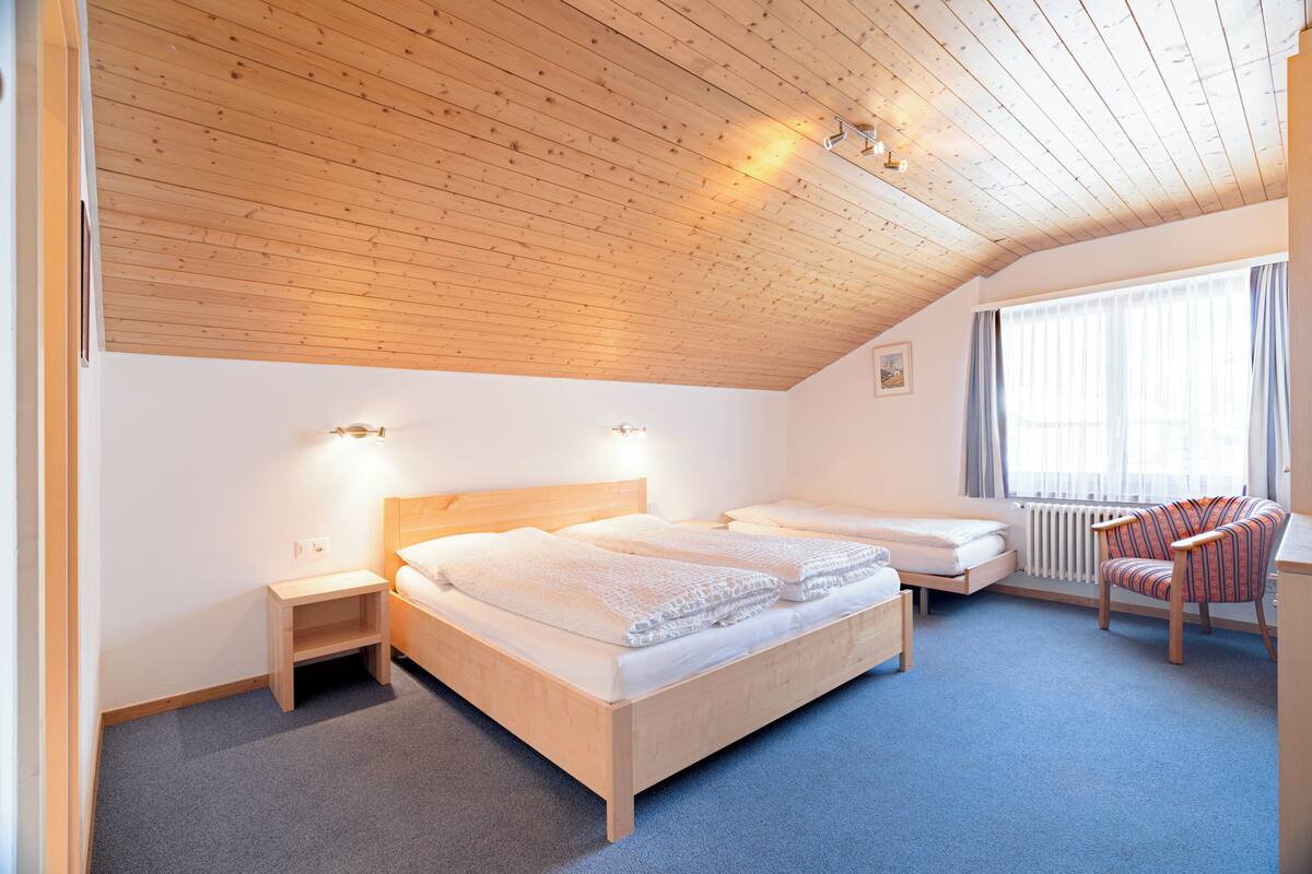 Quarto com duas camas, teto de madeira, janela com cortinas e uma cadeira.