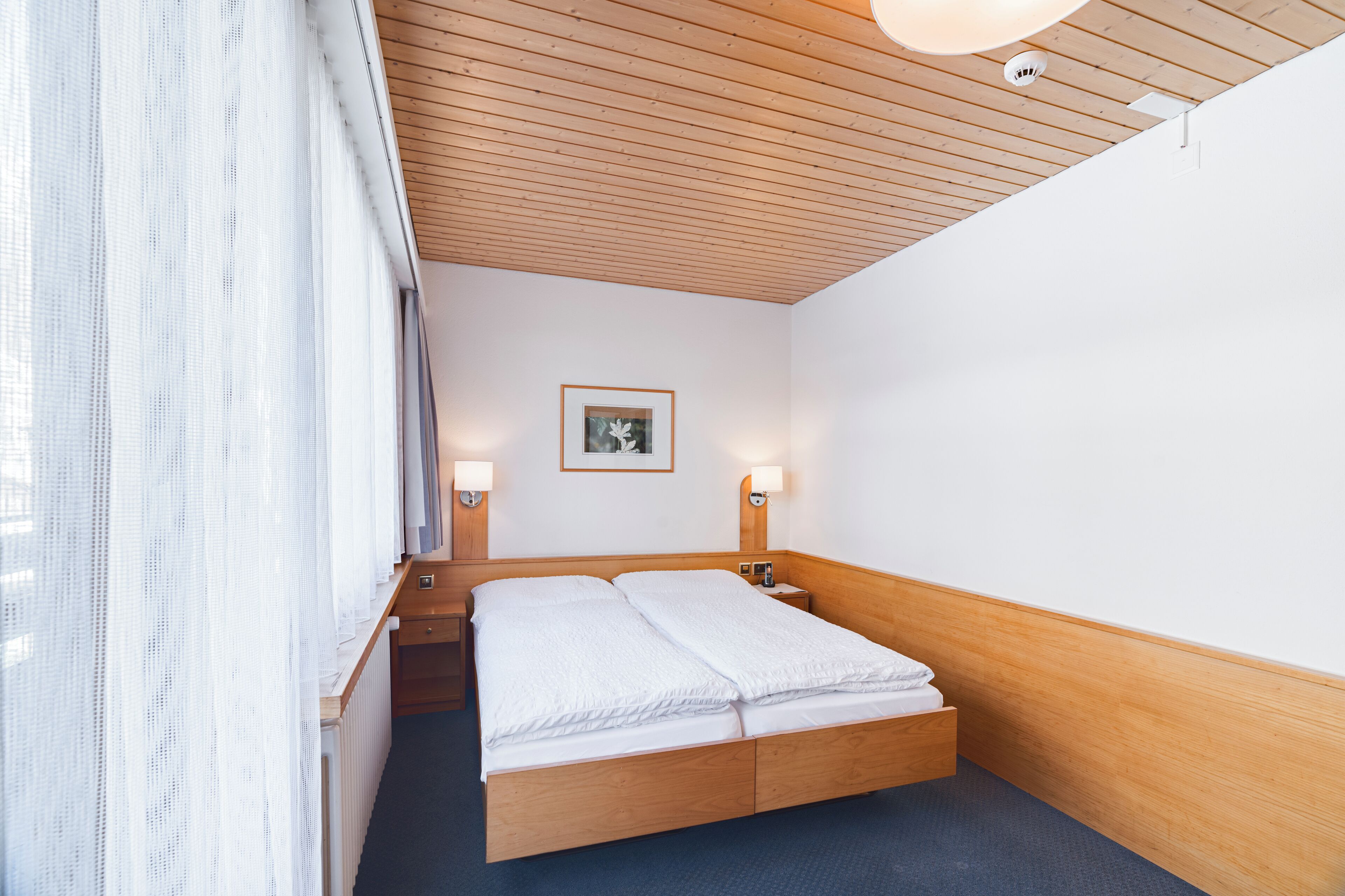 A hotel room with a wooden headboard, a double bed, two bedside lamps, a framed picture on the wall, and a window with sheer curtains.