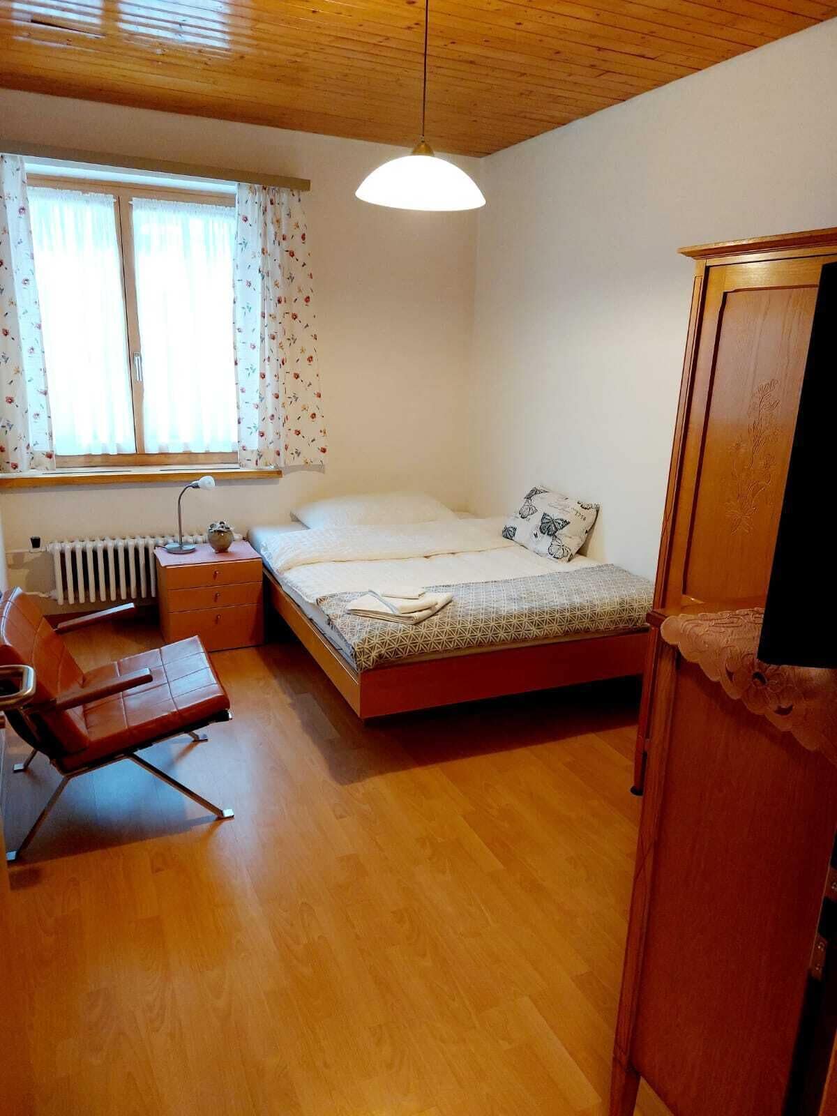 A bedroom with a wooden ceiling, a bed with patterned bedding, a chair, a desk, and a radiator.