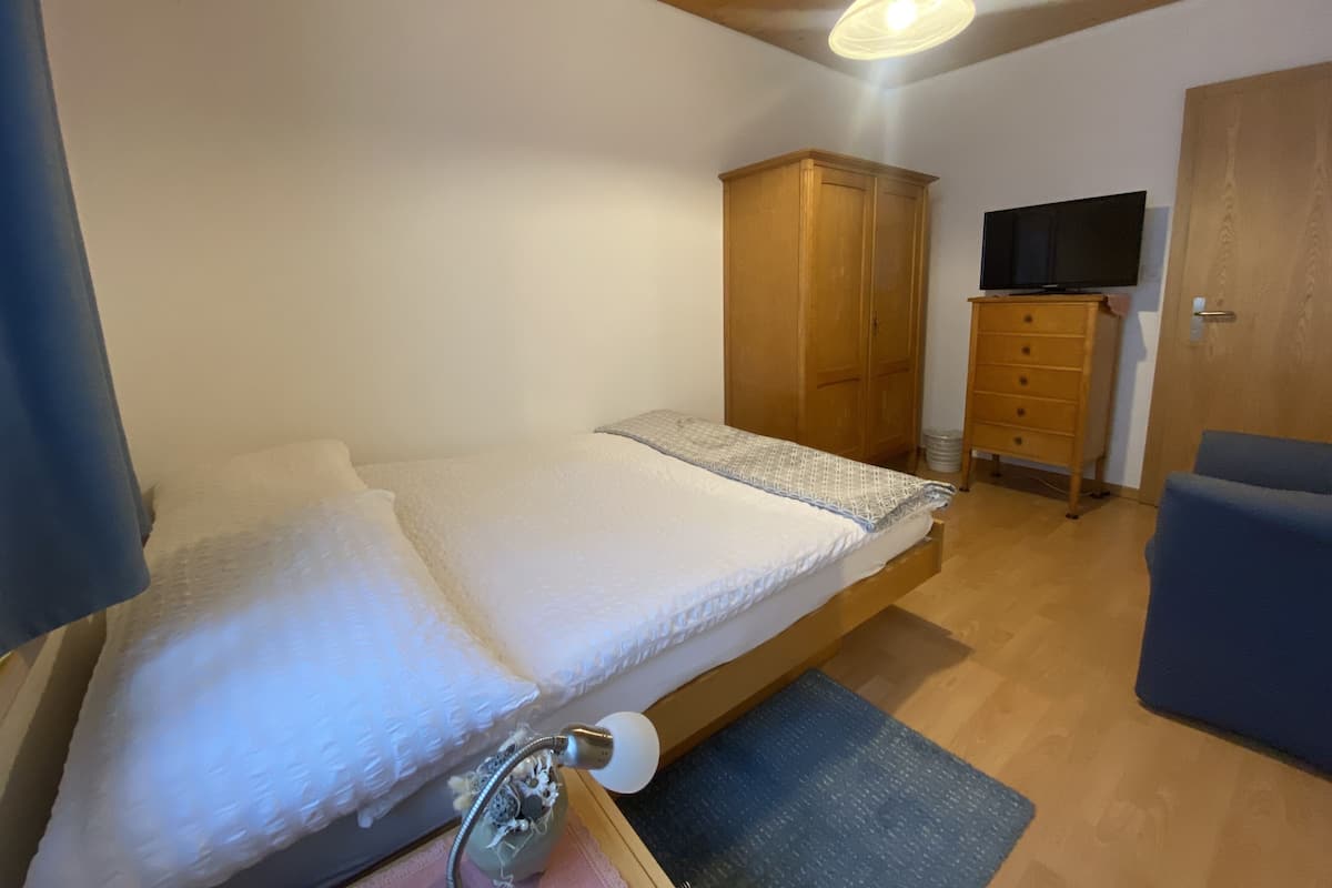 Quarto com cama, guarda-roupa de madeira, televisão e uma cadeira azul.