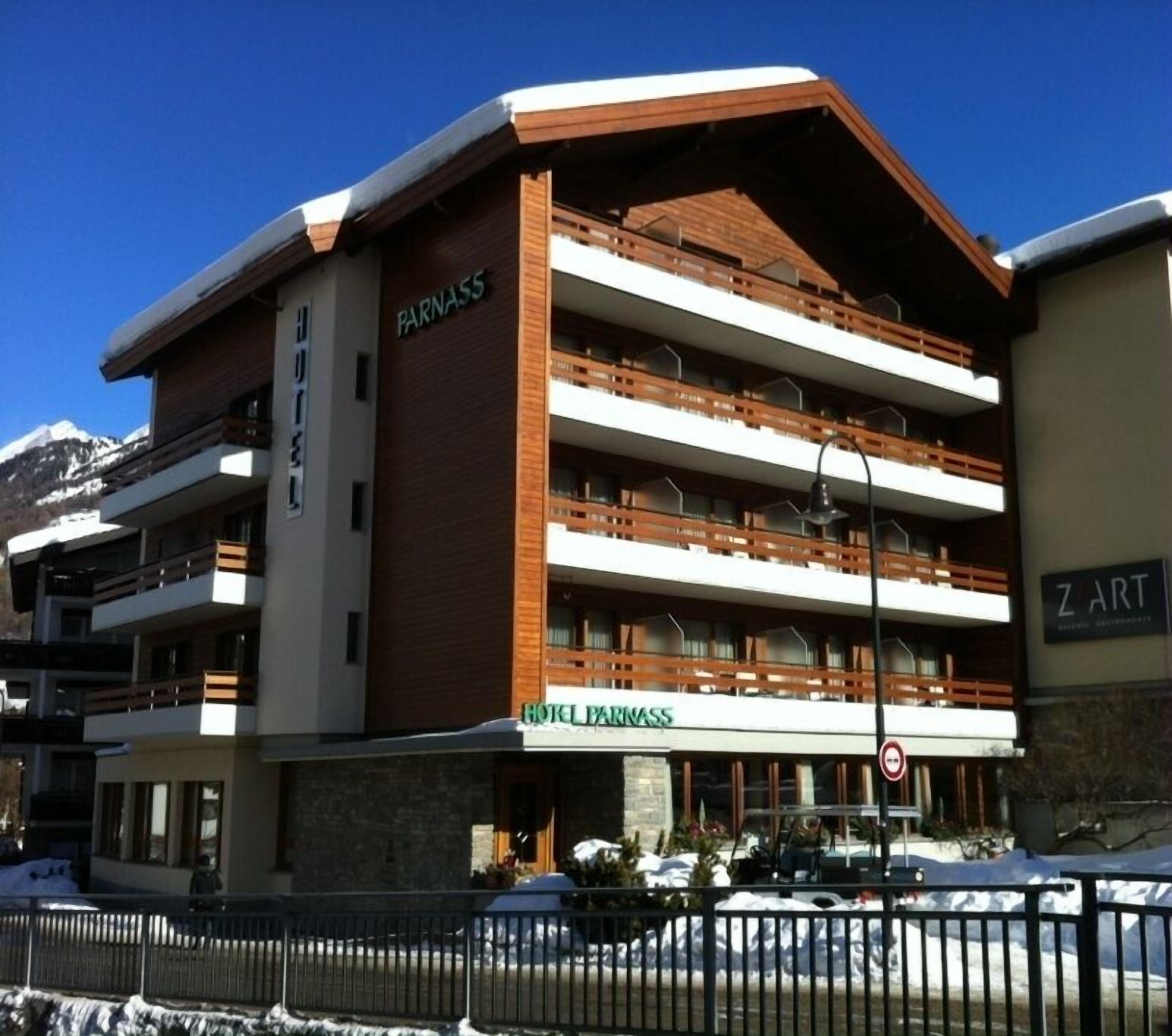 Een meerlaags hotel met een opvallend bord waarop 'HOTEL PARNASS' staat. Het heeft balkons op elke verdieping en een met sneeuw bedekte grond.