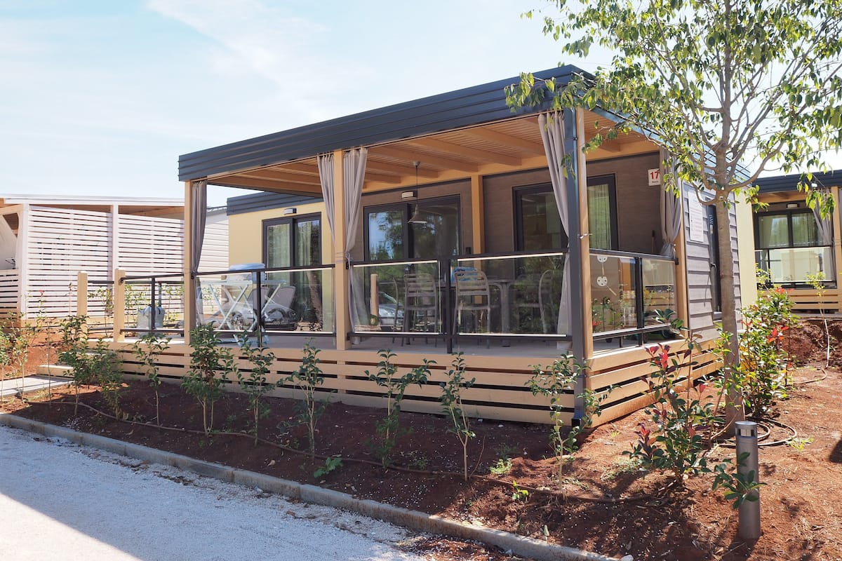 Une maison mobile moderne dotée d’un patio couvert, de grandes fenêtres et d’un jardin agrémenté de jeunes plantes.