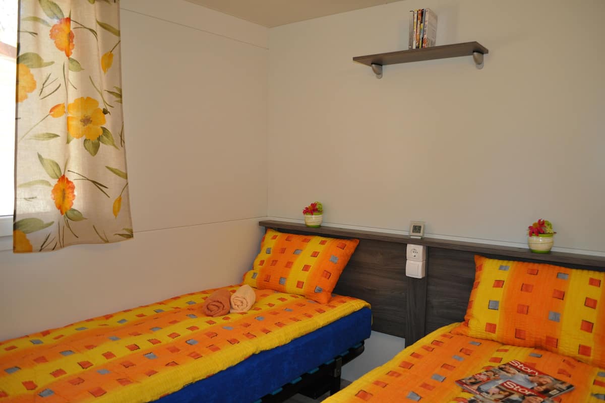 Une chambre avec deux lits, chacun agrémenté d’une couvre-lit colorée et de coussins assortis. Une étagère au-dessus des lits contient des livres et une petite plante.