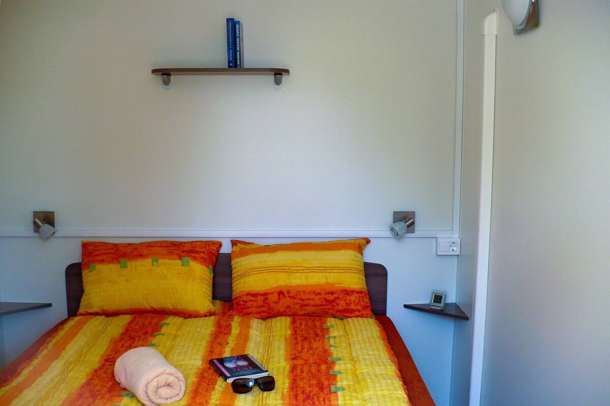 Un lit avec une couvre-lit à rayures, deux oreillers, une serviette pliée et un livre sur lequel reposent des lunettes de soleil.