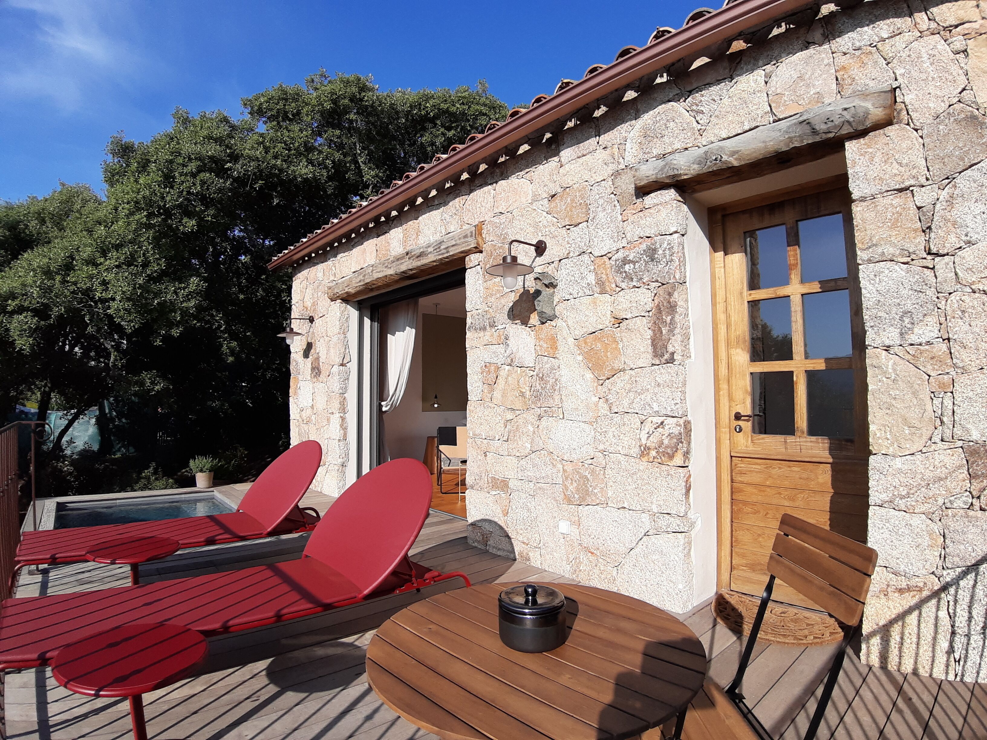 Un edificio de piedra con puerta y ventanas de madera, un área de piscina con sillas reclinables rojas y una mesa, y vistas a árboles al fondo.