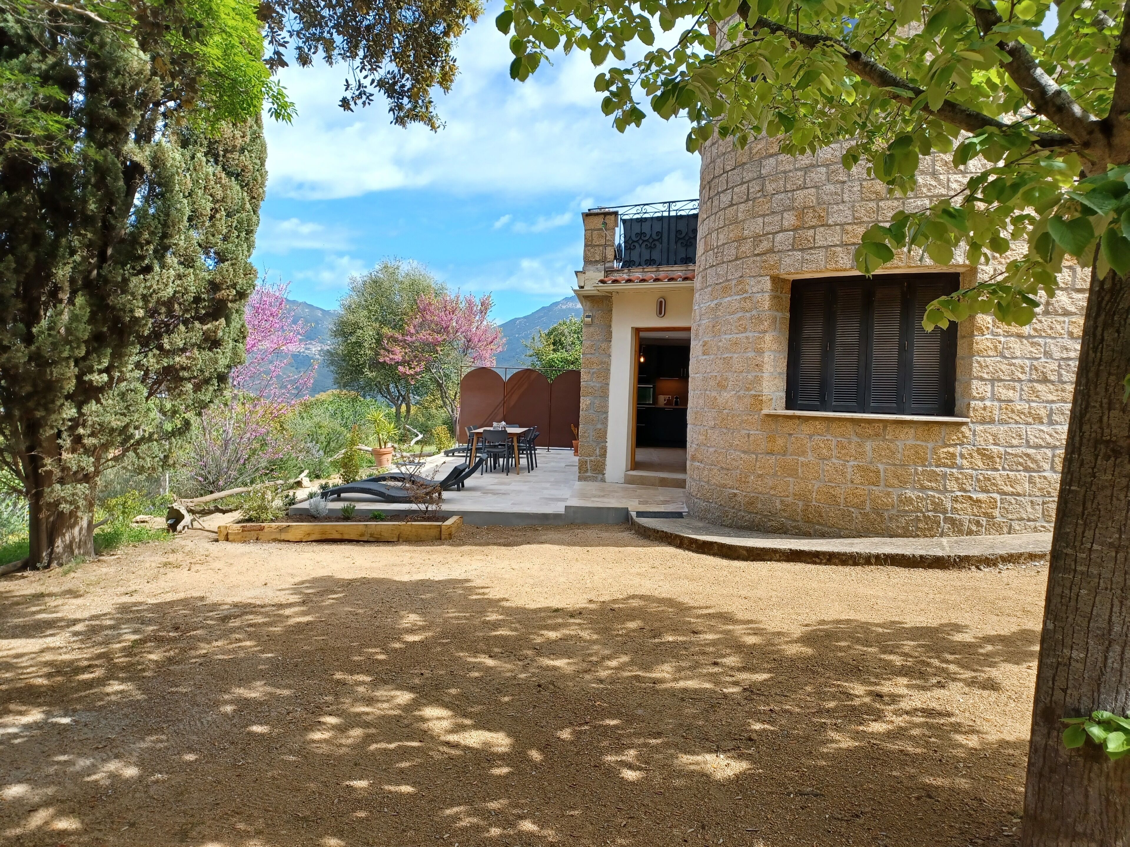Ein Steinhaus mit Balkon, einer Terrasse mit Tisch und Stühlen sowie einem Garten mit Bäumen und einer hölzernen Bank.