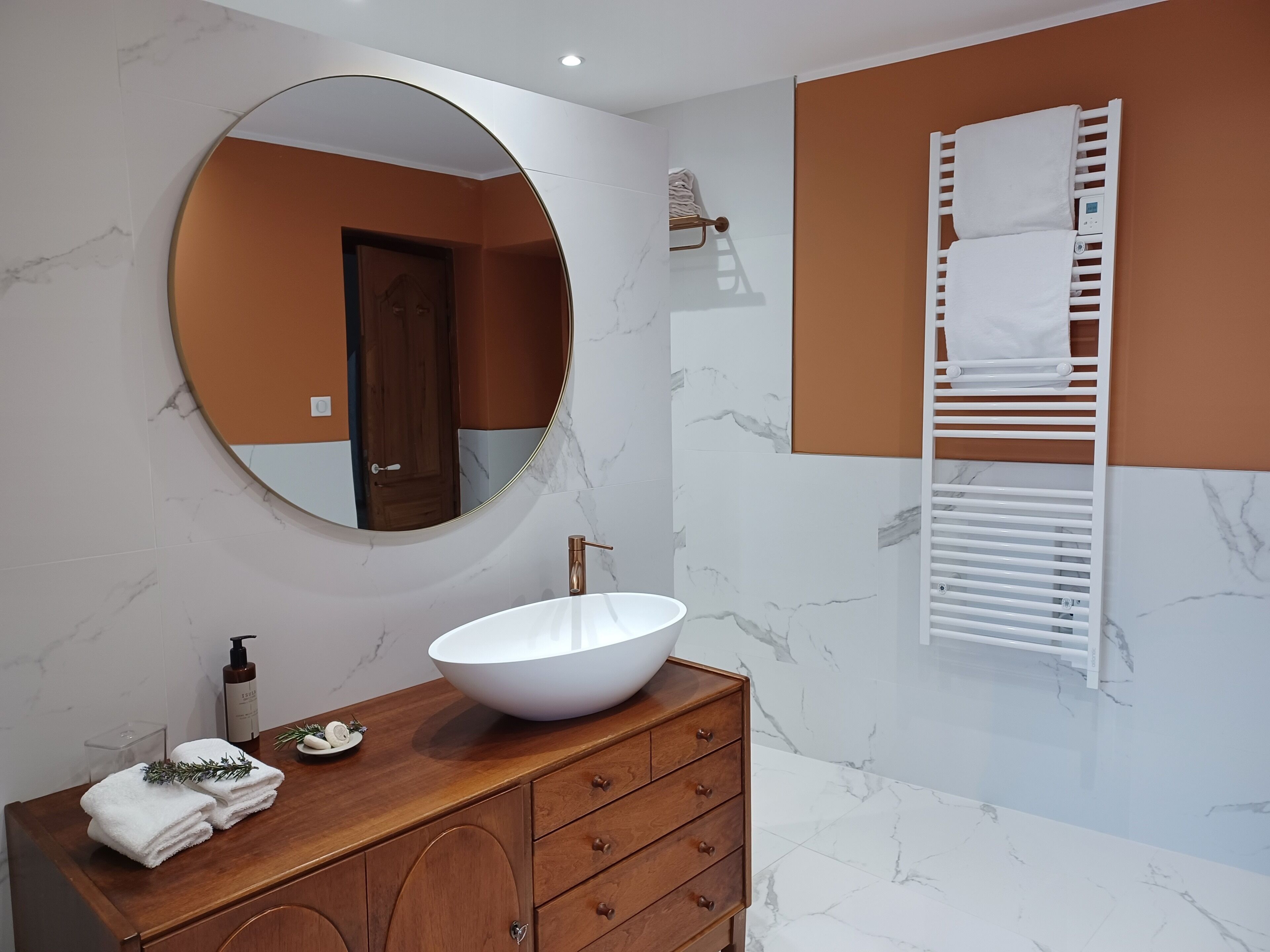 Une salle de bain moderne avec un miroir rond, un lavabo sur colonne blanc, un meuble-lavabo en bois et un porte-serviettes.